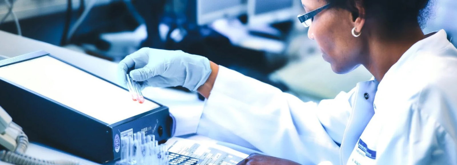 Person in weißem Laborkittel und Handschuhen hält eine Pipette über Reagenzgläser in einem Labor