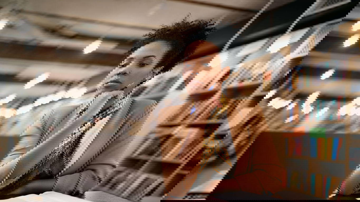 Alternative-Karrierewege.jpg Eine afroamerikanische Frau sitzt mit nachdenklicher Miene in einer Bibliothek vor dem Laptop