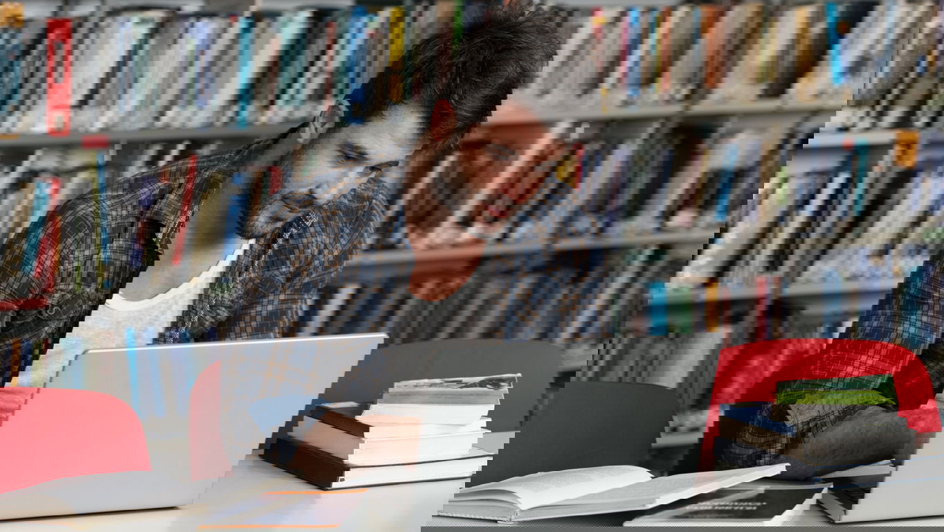 dissertationsthema.jpg Ein junger Mann schreibt in der Bibliothek an seiner Doktorarbeit