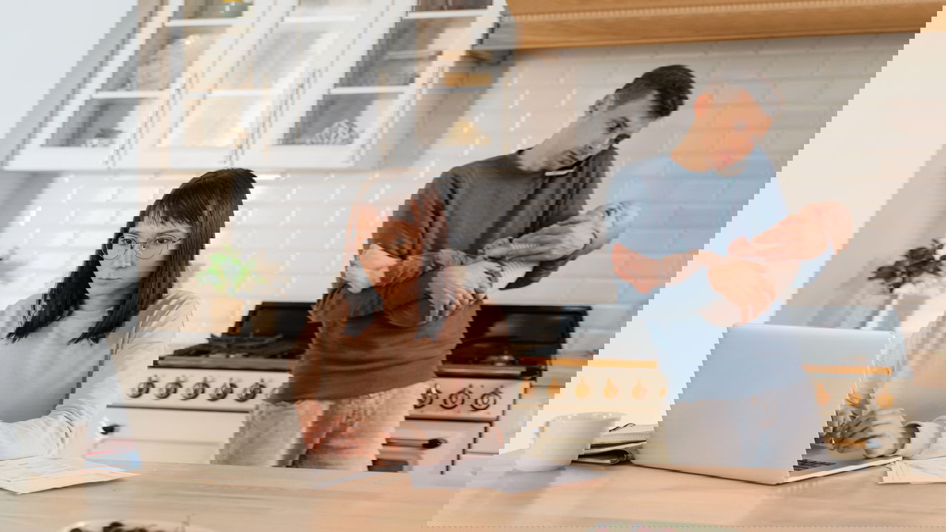 Forschung-Familie1.jpg Job und Familie: Eine Frau am Laptop und ein Mann mit Handy am Ohr und Kind auf dem Arm in der Küche