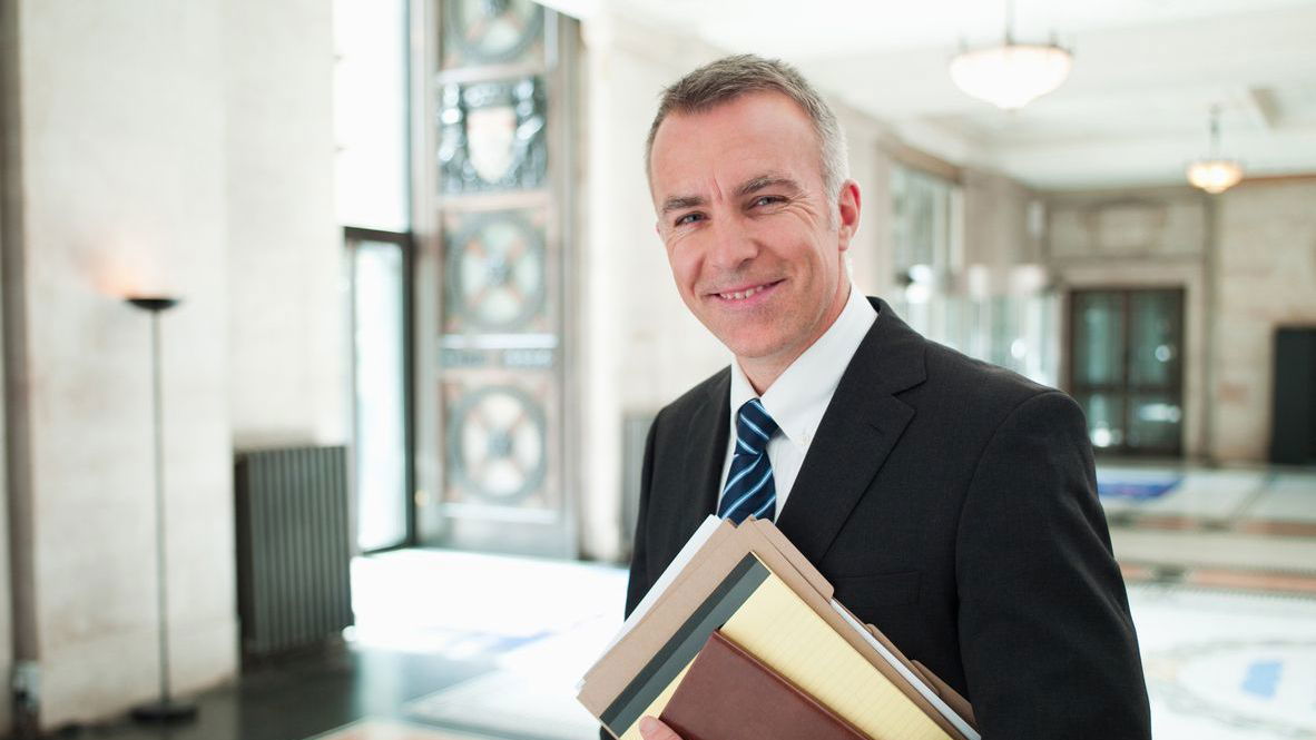 jurist-gehalt.jpg Ein lächelnder, älterer, weißer Jurist in einer Lobby mit Akten unter dem Arm