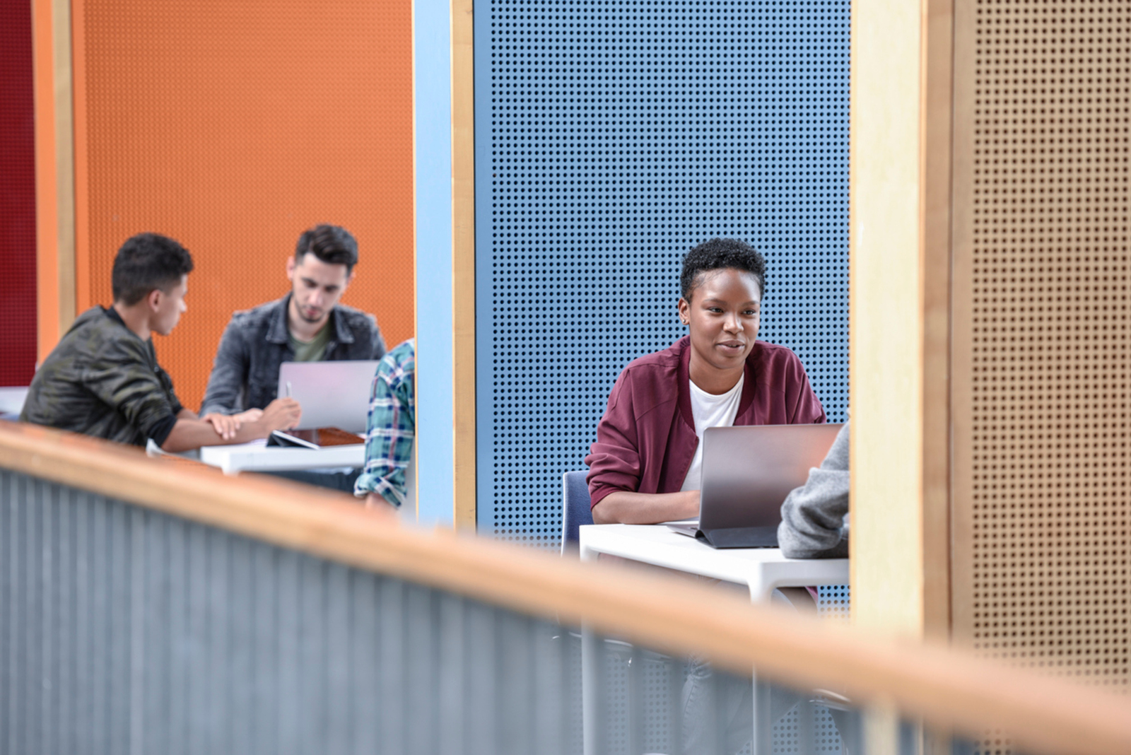 Mehrere Studierende sitzen mit Laptops an Tischen in einem modernen Innenraum, getrennt durch orange/blaue Wände.