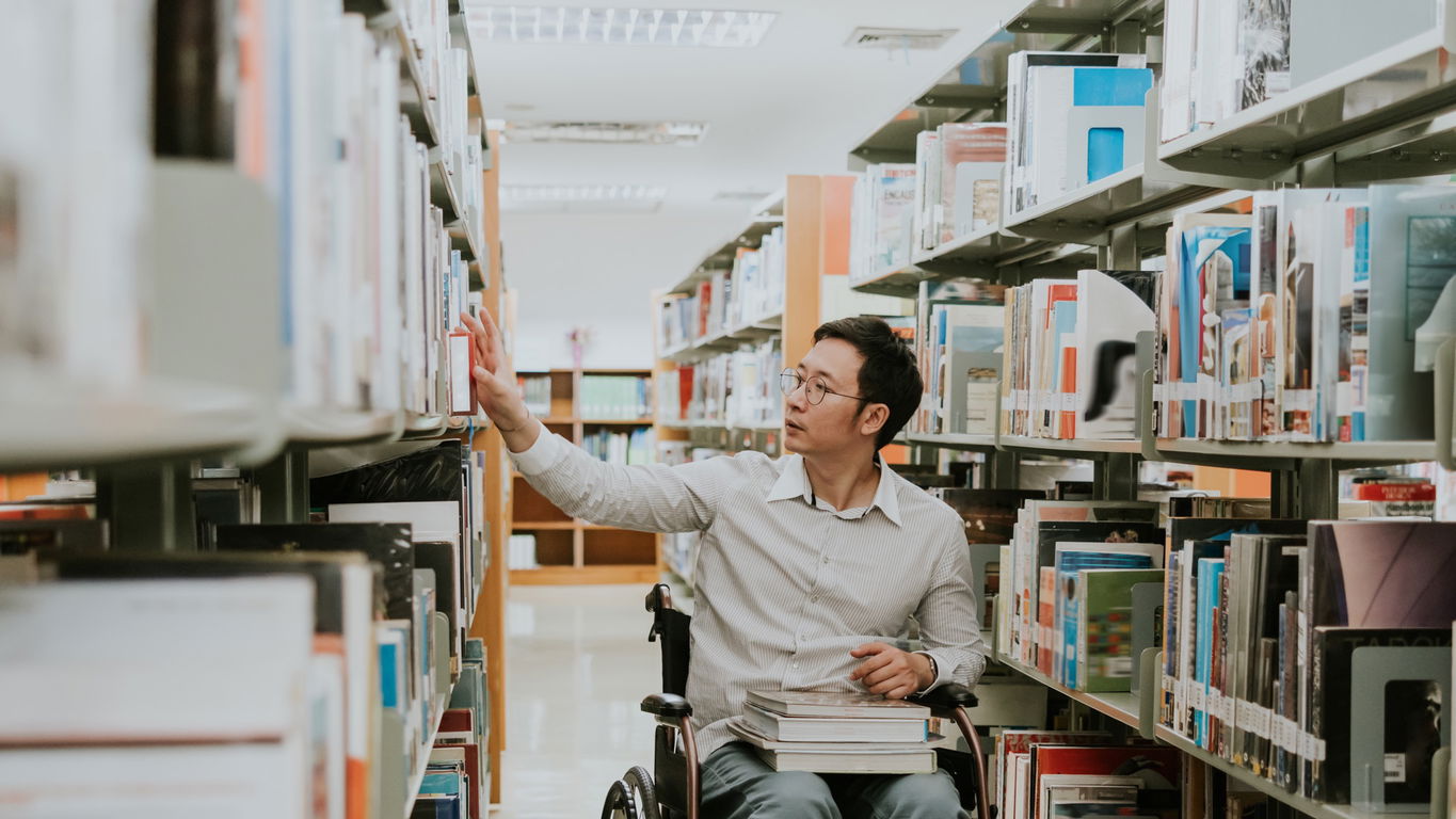 promovieren-mit-beeintraechtigung.jpg Ein asiatisch aussehender Doktorand im Rollstuhl in einer Bibliothek
