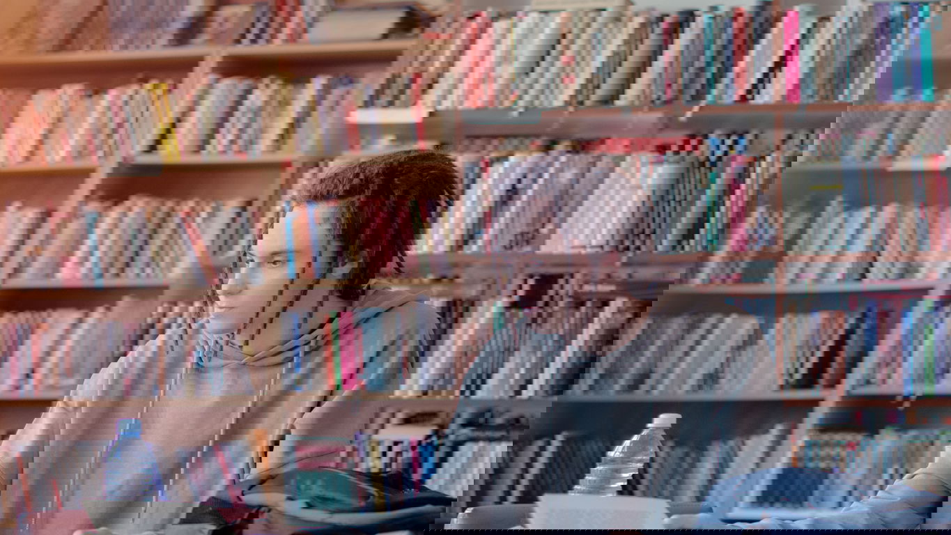 wissenschaftlicher-mitarbeiter-gehalt.jpg Ein Wissenschaftlicher Mitarbeiter mit dunklen Dreadlocks an einem Schreibtisch in der Bibliothek