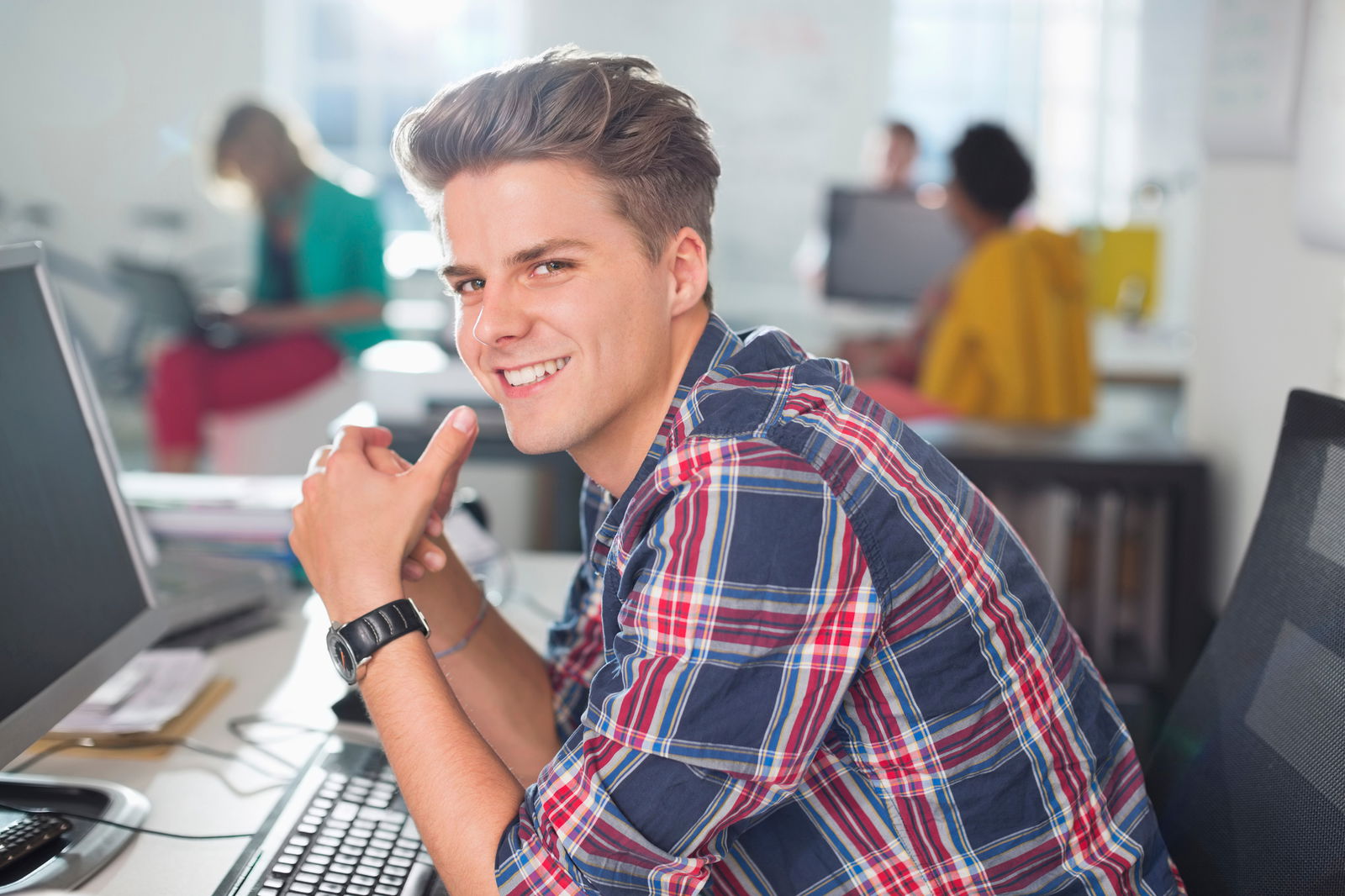 Ein lächelnder wissenschaftlicher Mitarbeiter in kariertem Hemd sitzt vor dem Computer am Schreibtisch