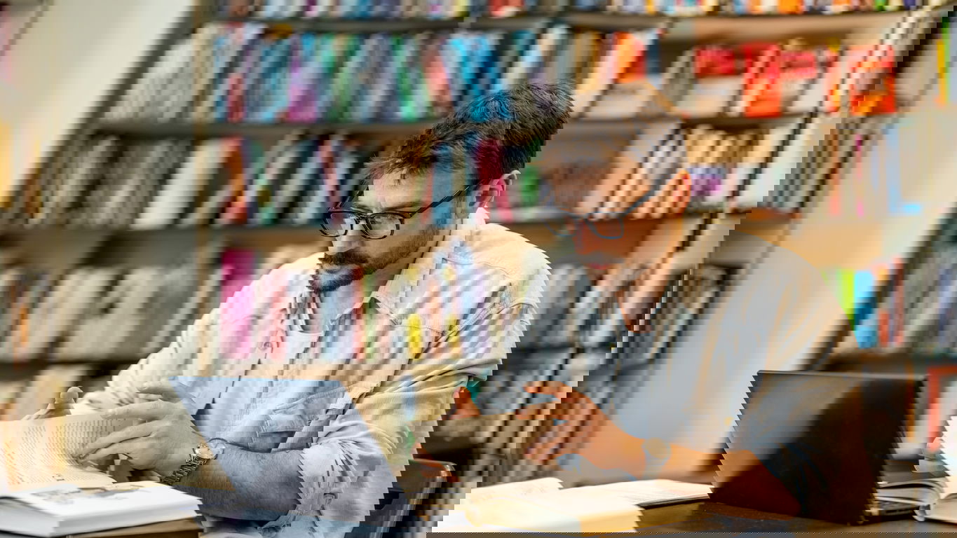 zitierregeln.jpg Ein junger Mann am Laptop in einer Bibliothek will aus einem Buch zitieren